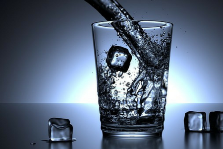 Vatten hälls i ett glas. Iskuber är både i glaset och på bordet som glaset står på.