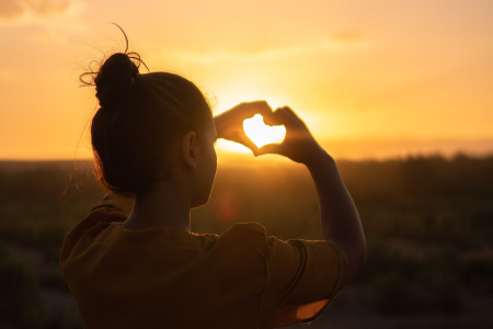 Kvinna sitter i motljus av solnedgång och formar händerna till ett hjärta