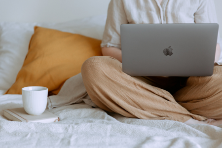 Anonym person sitter i en säng med en dator i knät och en mugg bredvid.