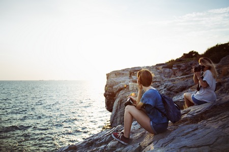 Två personer med kameror sitter på klippor vid havet.