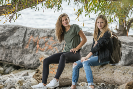 Två tonårsflickor sitter på en sten. Ett träds grenar syns ovanför dem och i bakgrunden syns vågor på en sjö.