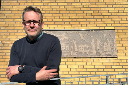 Ny fastighetschef Per Olofsson står ute i solen framför gul tegelvägg med armarna i kors.